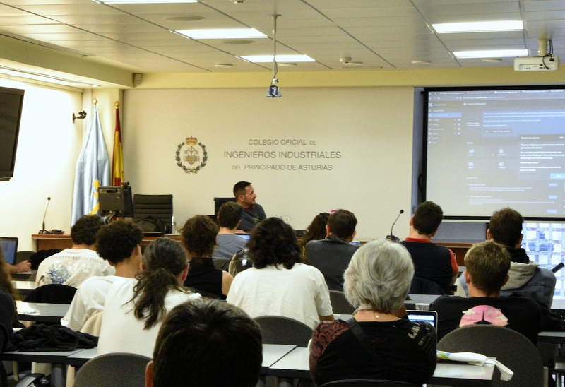 A group of people sit at tables in a presentation hall. There is a projector screen at the front of the room, with a logo for "Colegio Oficial de Ingenieros Industriales del Principado de Asturias" above it. The people in the room are mostly students, and they look like they're paying attention to the presentation.